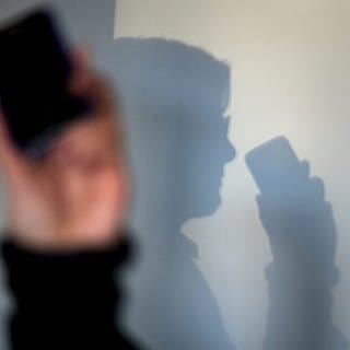 Eine Hand hält ein Mobiltelefon. An der Wand dahinter ist der zugehörige Schatten eines Mannes zu sehen.